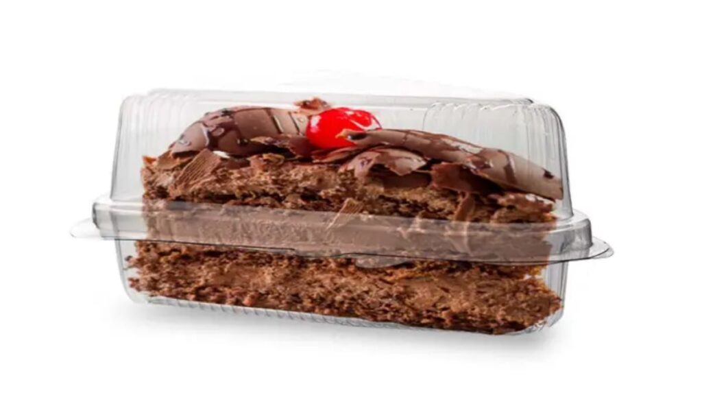 Fatia de bolo de chocolate com cereja em uma embalagem descartável transparente.
